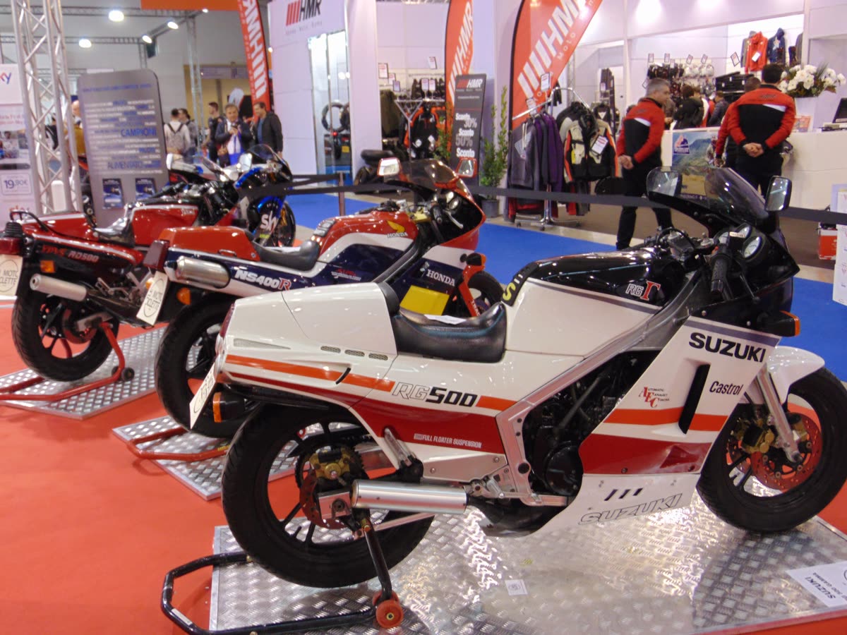 Suzuki RG500 Gamma blanche et rouge exposée dans un salon moto, à côté d'une Honda NS400R